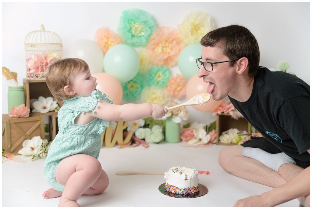 baby girl feeding her dad cake with wooden spoon and peach and mint decorations for cake smash 